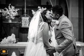 Inside a Melbourne church in Victoria, Australia, a black and white image shows the priest standing behind the couple, discreetly watching as they share their first kiss, adding a lighthearted and candid touch to the ceremony.